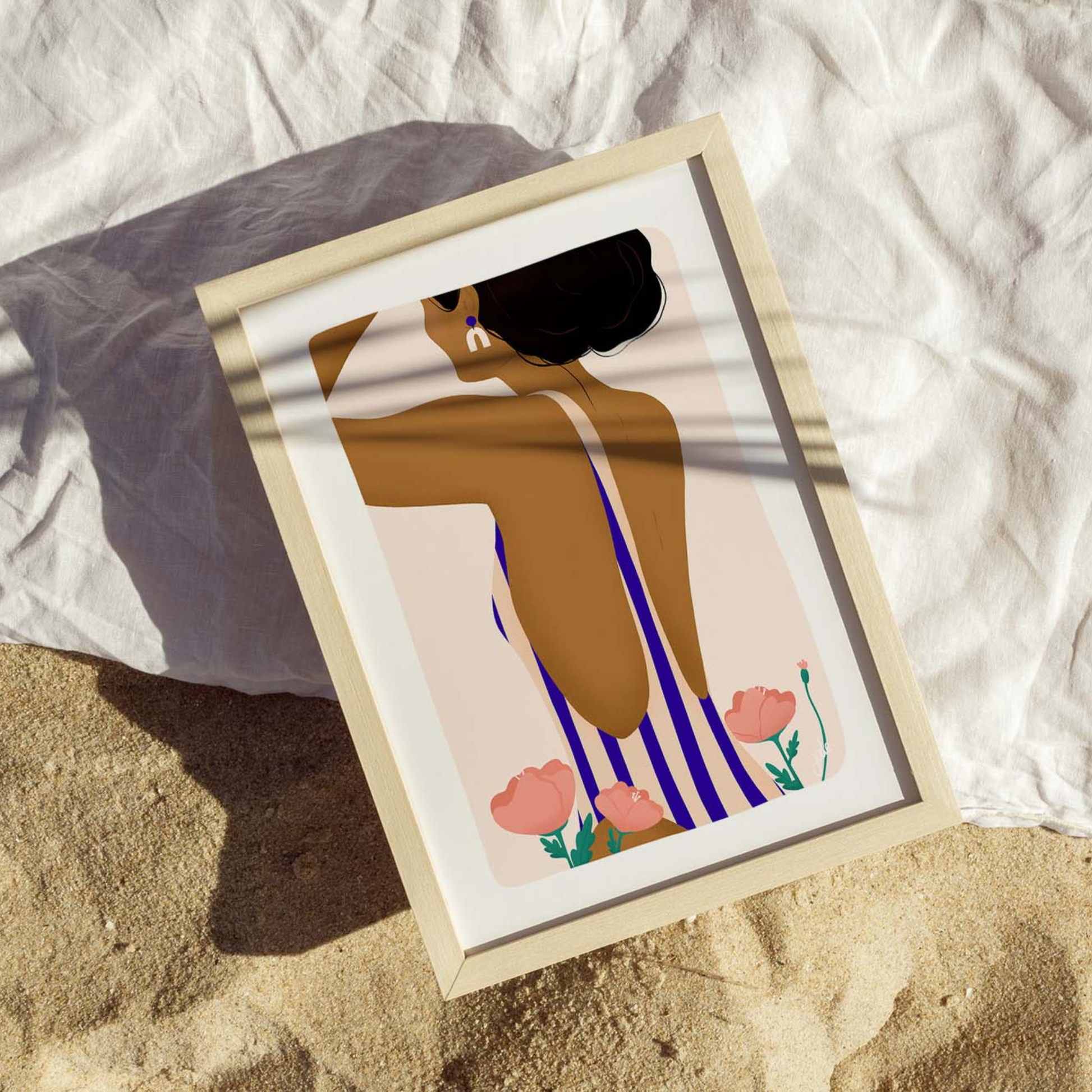 Une illustration encadrée représentant une silhouette féminine de dos est posée sur un tissu clair dans un décor sableux.