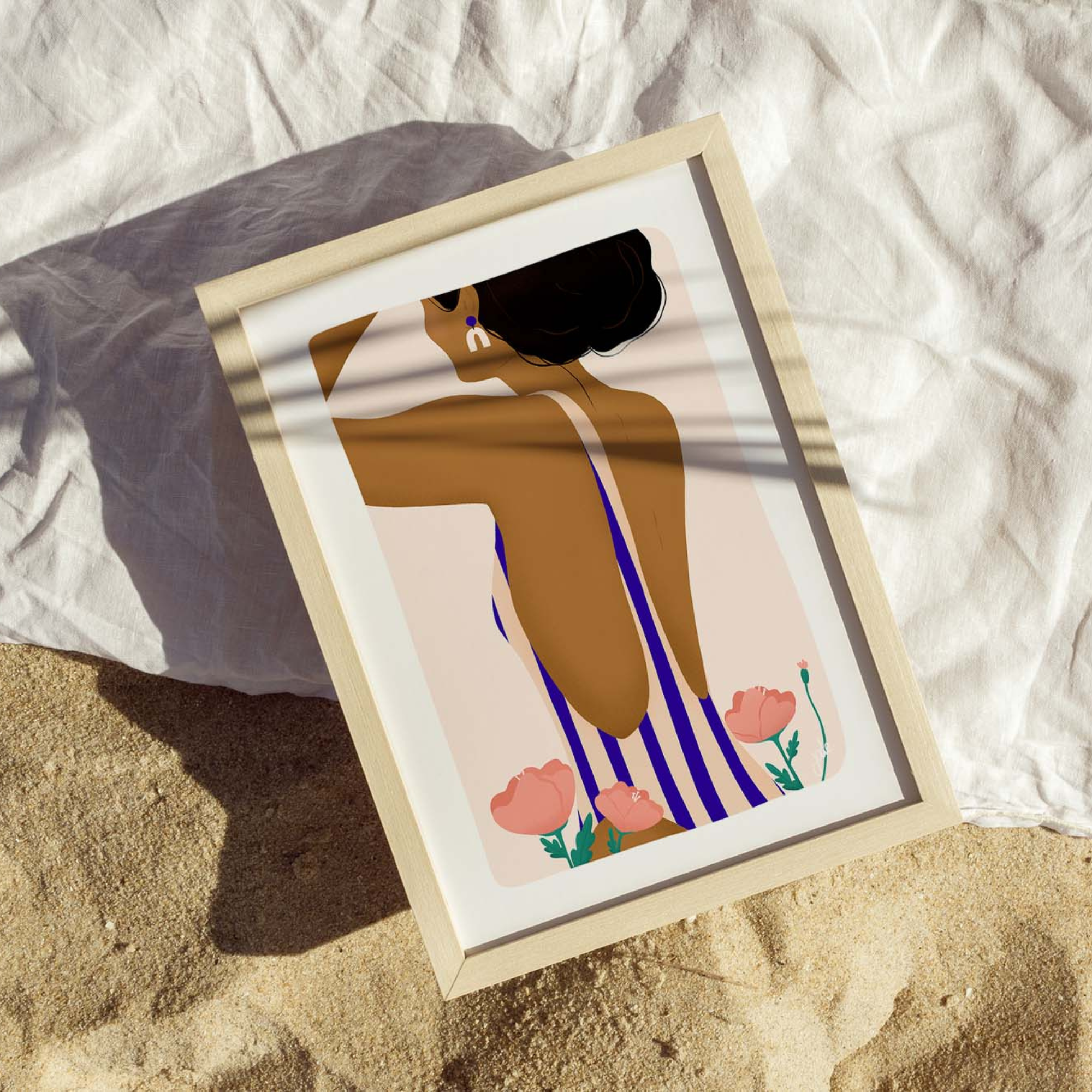 Une illustration encadrée représentant une silhouette féminine de dos est posée sur un tissu clair dans un décor sableux.