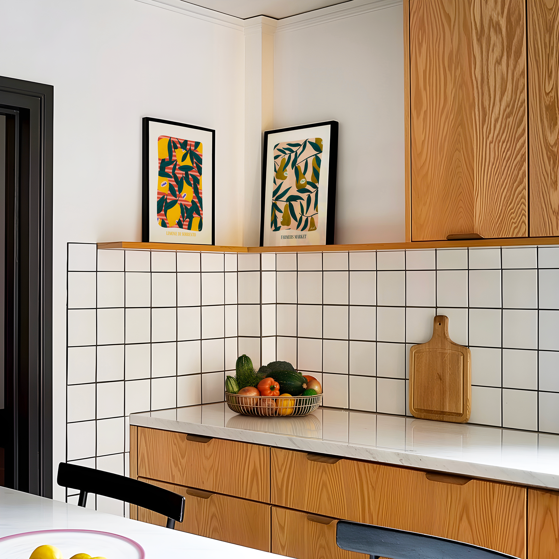 Deux affiches illustrées aux motifs de fruits et feuillages encadrées et posées sur une étagère d’angle dans une cuisine lumineuse, au-dessus d’un plan de travail avec corbeille de légumes et planche en bois.