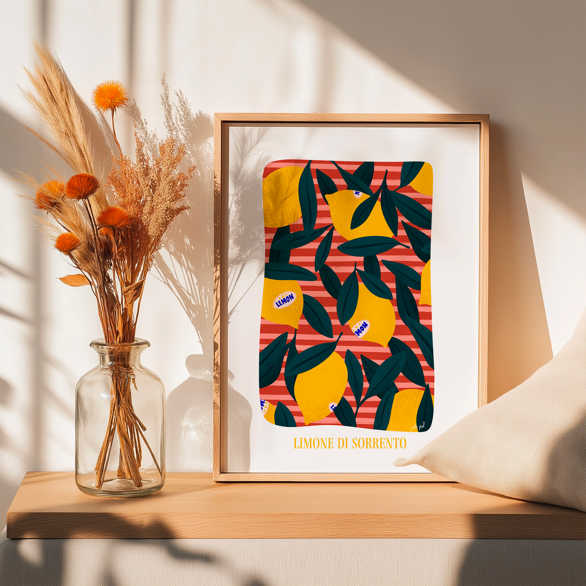 Affiche illustrée de citrons jaunes et feuillage vert sur fond rayé rouge, encadrée et posée sur une étagère en bois à côté d’un vase de fleurs séchées et d’un coussin beige, éclairée par une lumière naturelle.