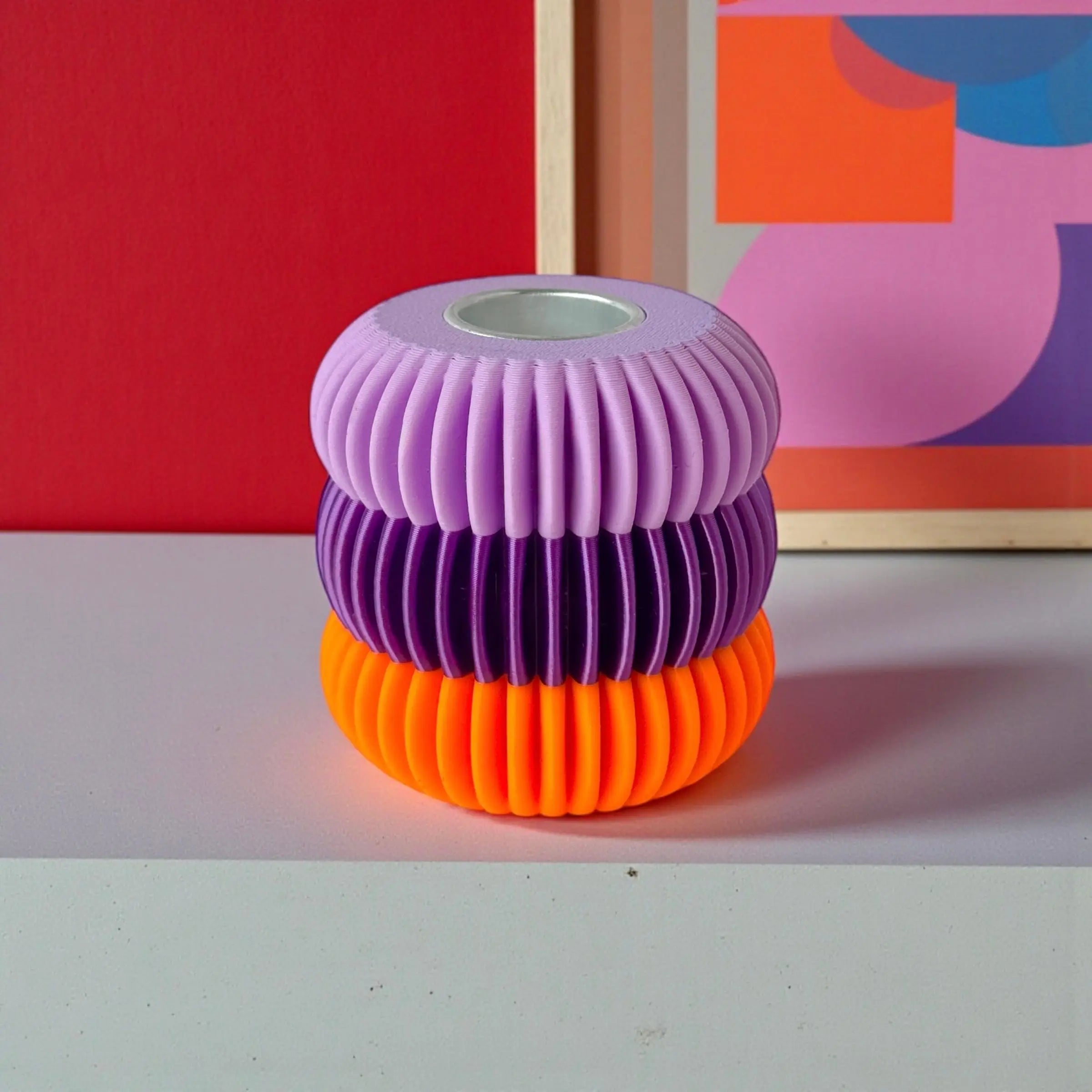 Bougeoir design nervuré en trois anneaux lilas, violet et orange, mis en scène dans un décor graphique et coloré aux accents pop.