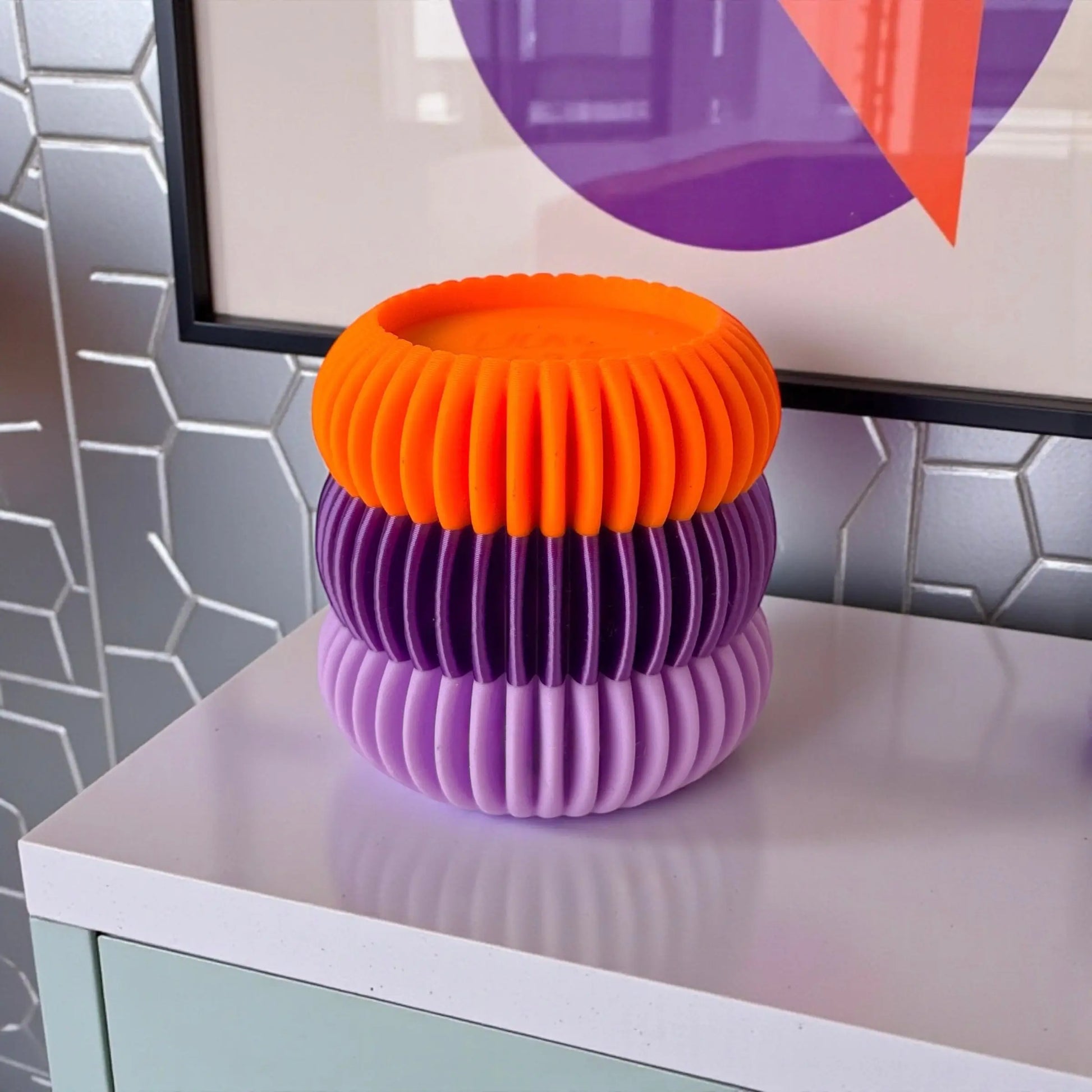 Bougeoir design nervuré en trois anneaux lilas, violet et orange, mis en scène dans un décor graphique et coloré aux accents pop.