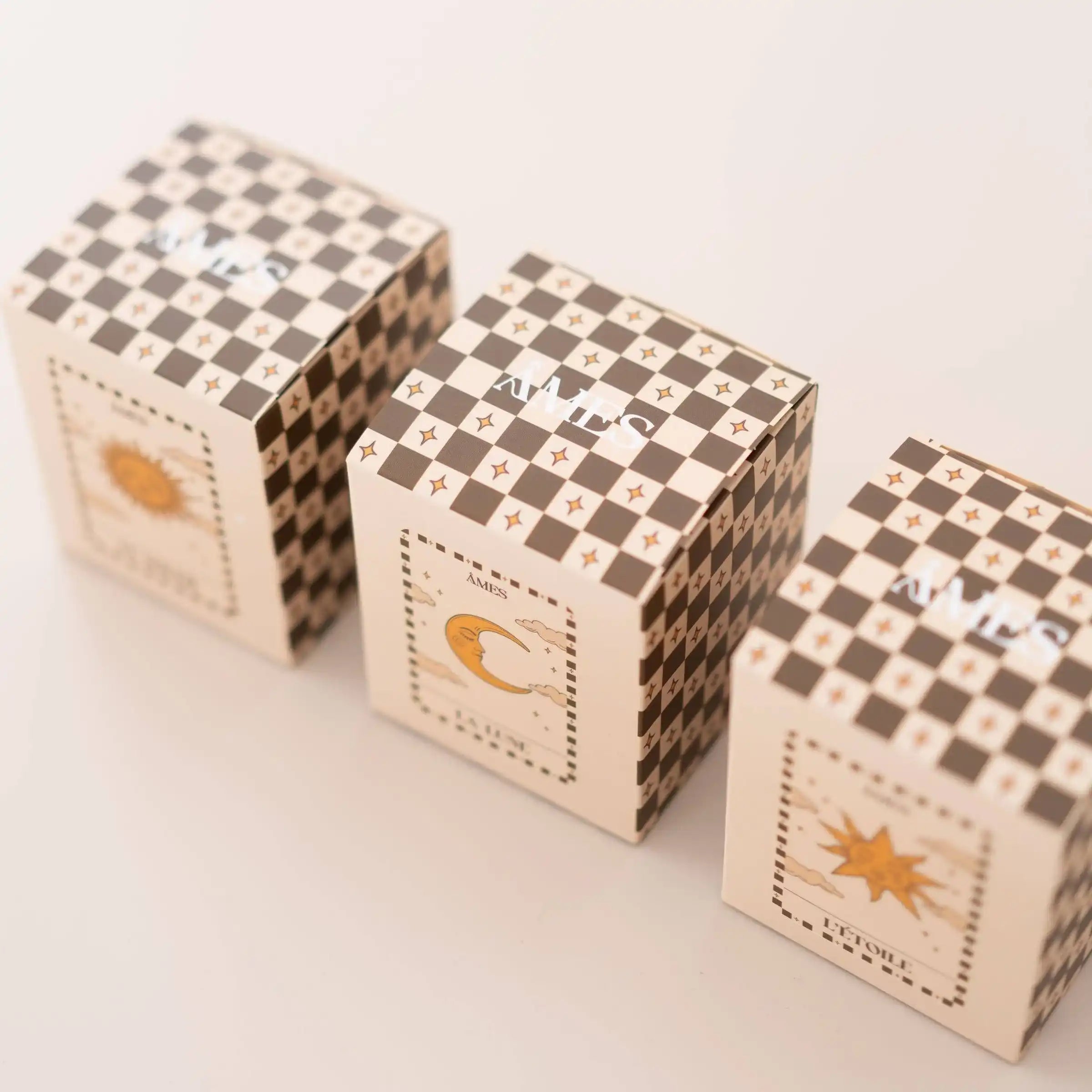 Trois coffrets de bougies parfumées AMES collection Tarot, alignés sur fond clair, avec un design damier élégant et des illustrations symboliques “Le Soleil”, “La Lune” et “L’Étoile”.