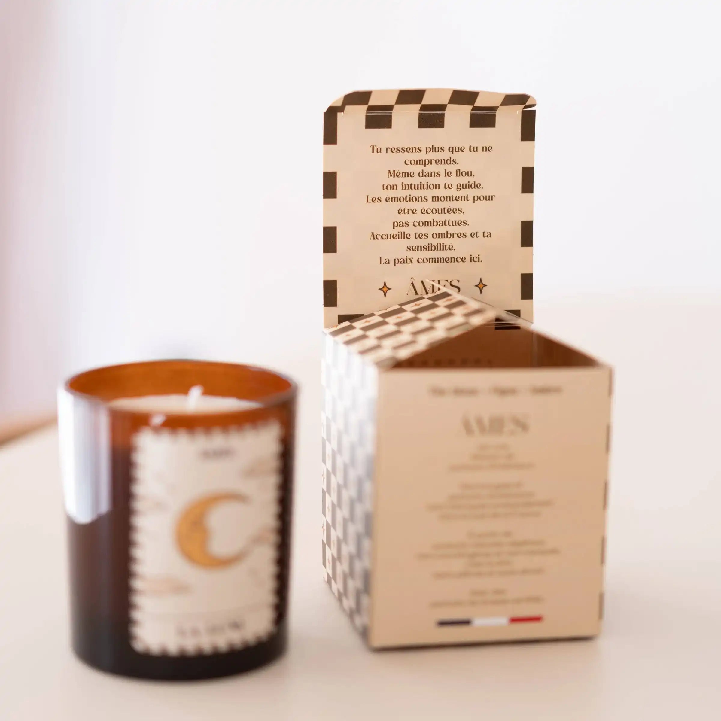 Une bougie parfumée AMES “La Lune” à côté de son coffret ouvert, laissant apparaître un message inspirant à l’intérieur du rabat.