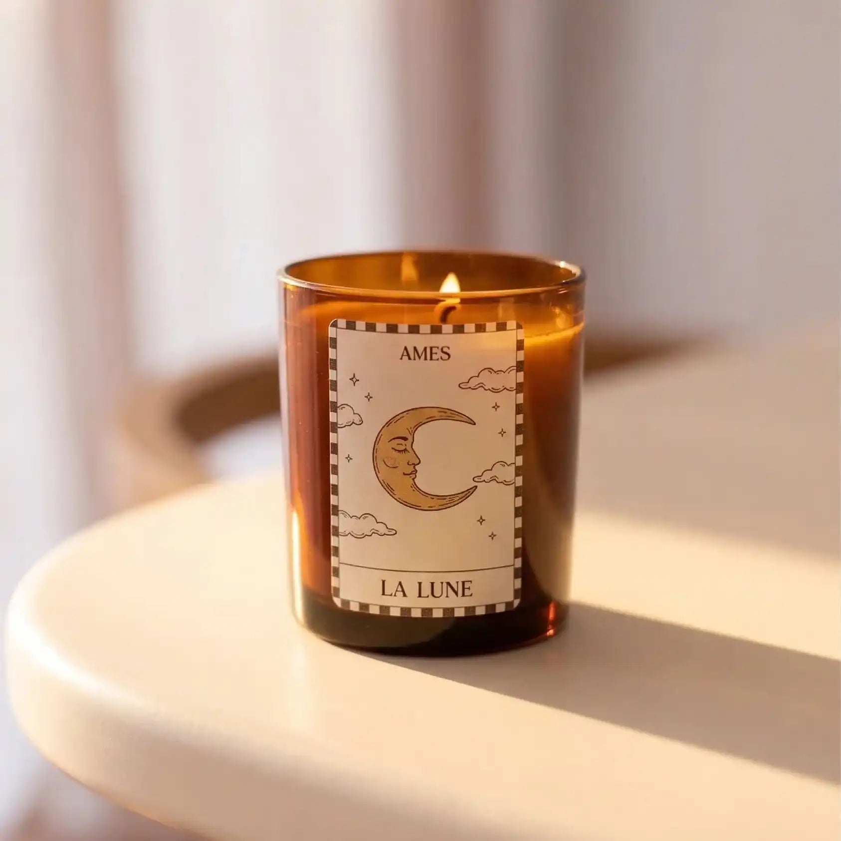 Bougie parfumée AMES “La Lune” allumée, présentée dans un élégant verre ambré avec une étiquette illustrée d’un croissant de lune, posée sur une table ronde baignée de lumière douce.
