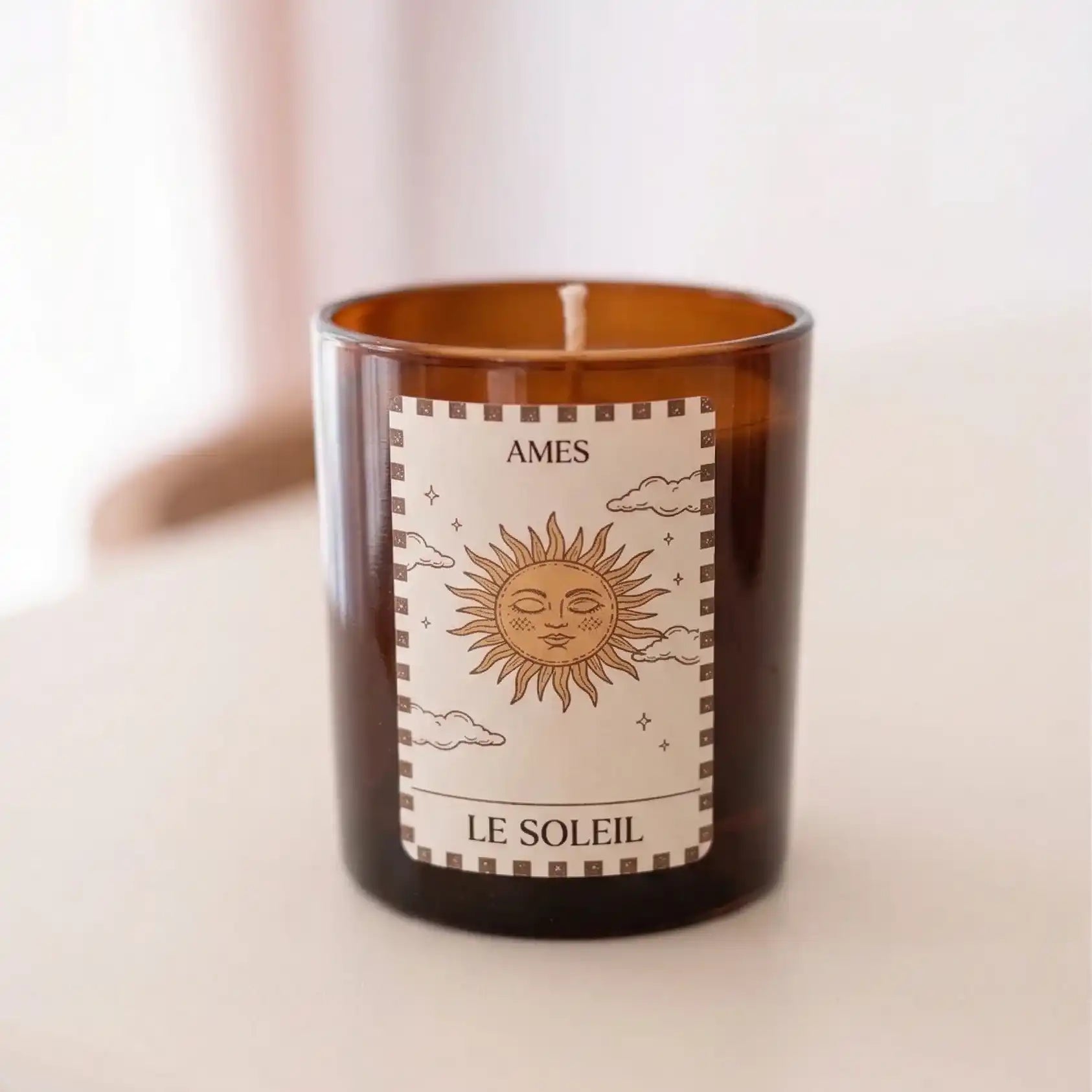 Bougie parfumée AMES “Le Soleil” dans un verre ambré, ornée d’une étiquette illustrée d’un soleil au style tarot, posée sur une table ronde dans une ambiance douce et lumineuse.