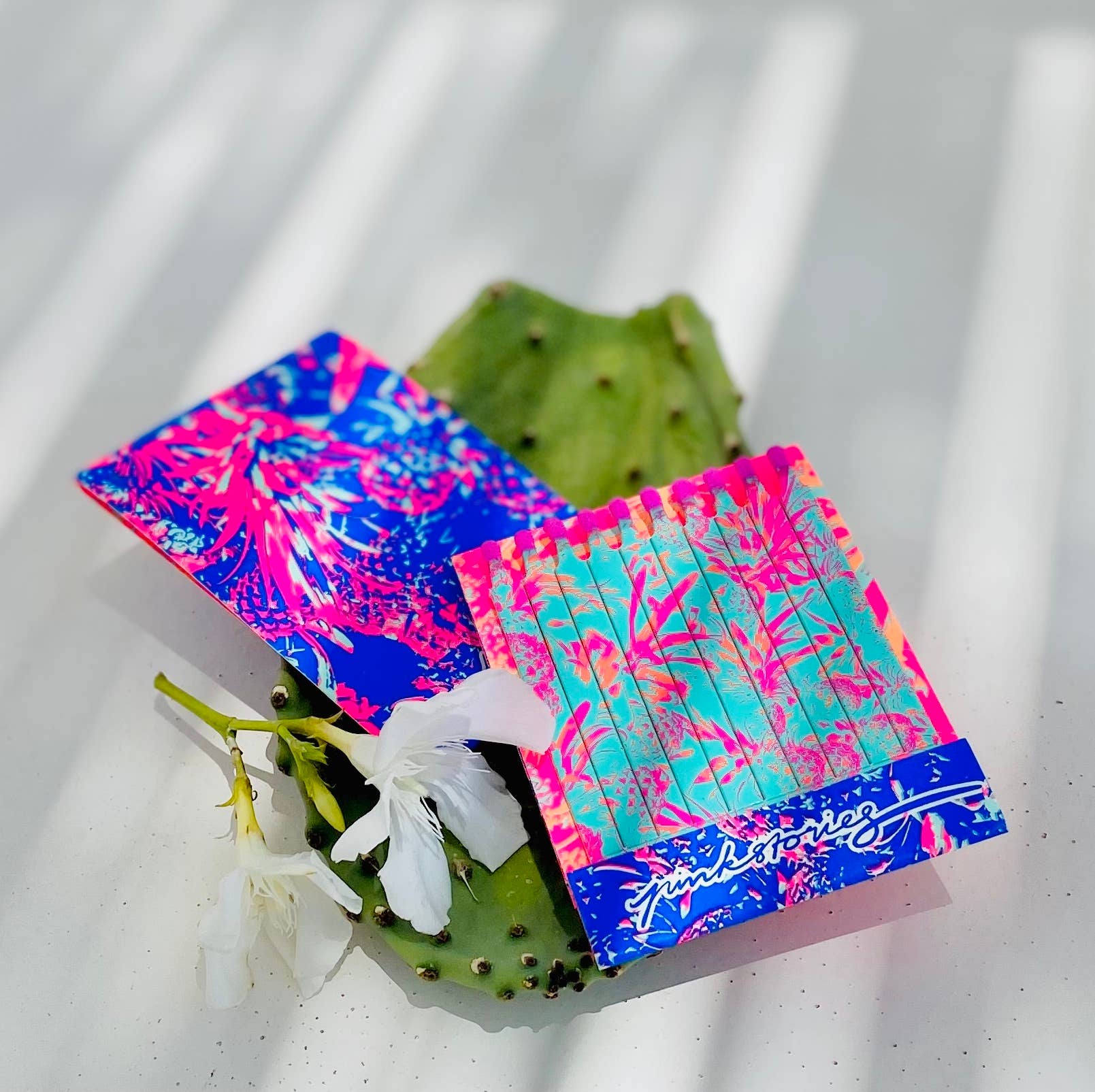 Carnet d’allumettes ouvert posé sur une feuille de cactus, avec design tropical turquoise, rose et bleu intense pour une mise en scène solaire et estivale.