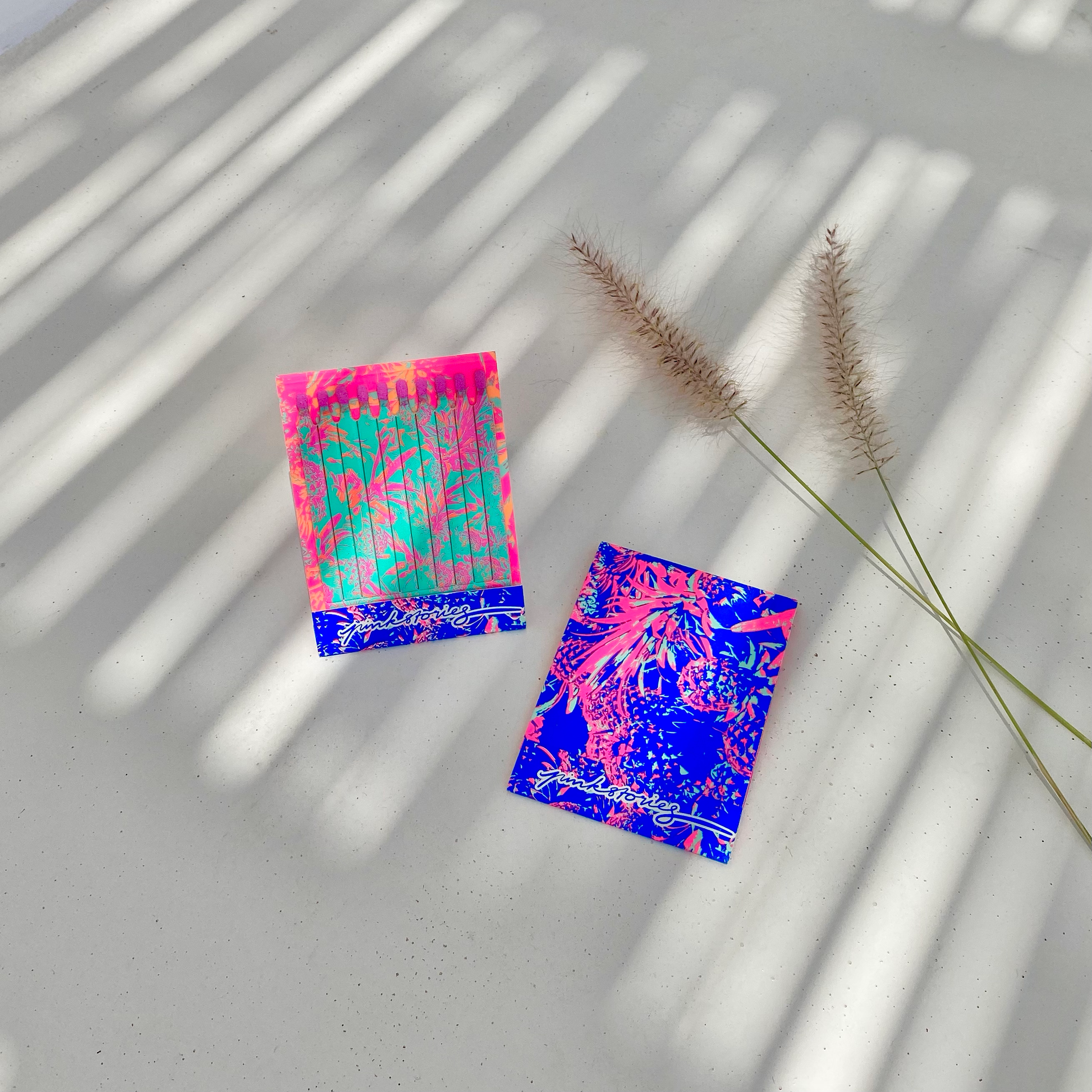 Deux carnets d’allumettes aux couleurs pop et tropicales, posés sur une surface claire avec un jeu d’ombres graphiques et quelques herbes séchées.