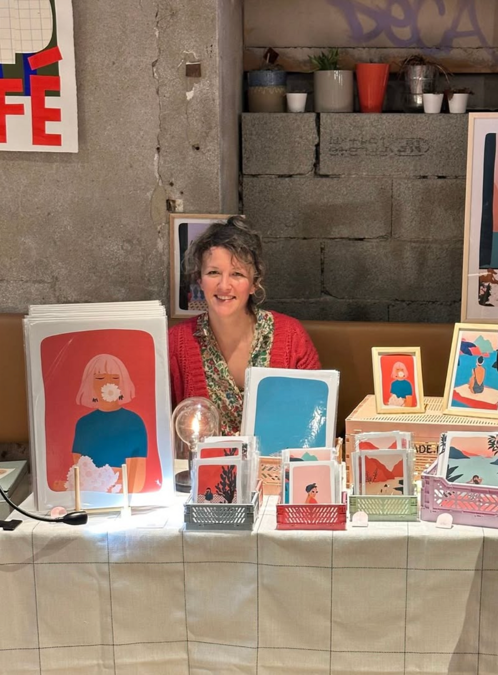 Illustratrice souriante présentant ses affiches colorées sur un stand d’exposition.