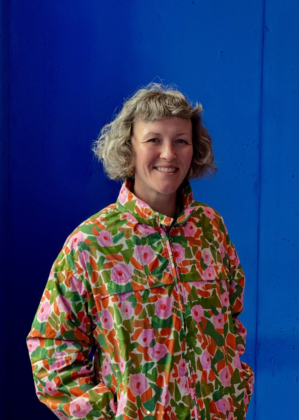 Portrait d’une femme souriante aux cheveux courts et bouclés, portant une veste fleurie devant un mur bleu vif.