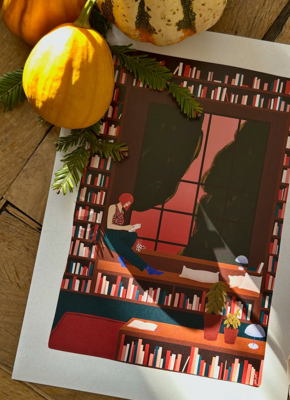 Illustration colorée posée sur une table en bois, représentant une femme assise dans une bibliothèque près d’une grande fenêtre, entourée de citrouilles décoratives et de branches de sapin.