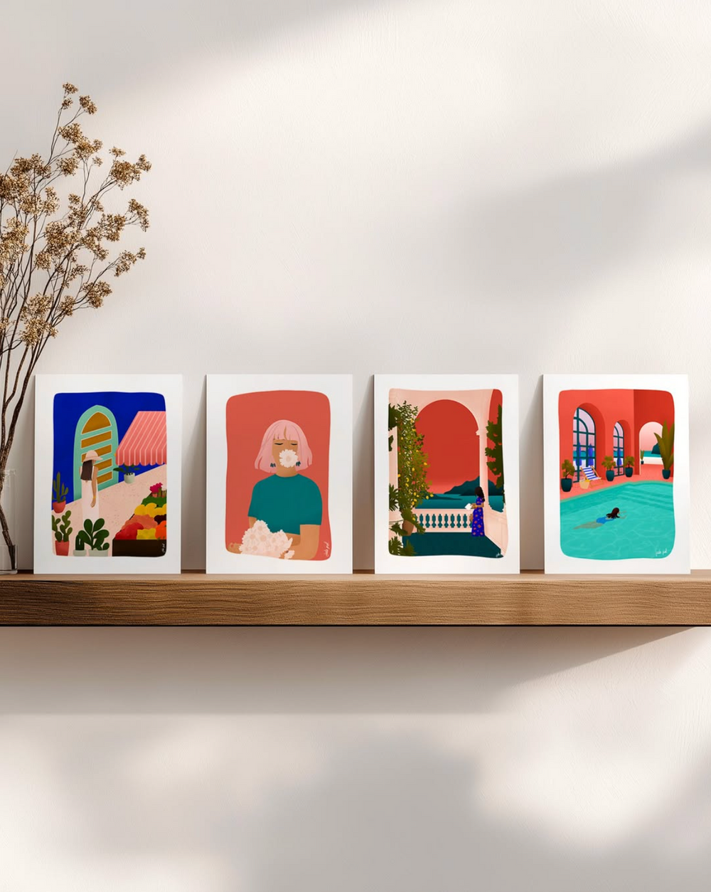 Quatre affiches illustrées aux couleurs vives sont posées côte à côte sur une étagère en bois contre un mur clair, accompagnées d’un bouquet de fleurs séchées.