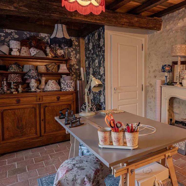 Atelier artisanal chaleureux installé dans une grange en pierre, rempli de lampes et abat-jour fleuris de styles variés, entre mobilier en bois, poutres apparentes et ambiance rétro.