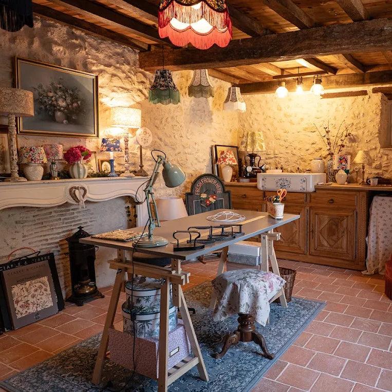 Atelier artisanal chaleureux installé dans une grange en pierre, rempli de lampes et abat-jour fleuris de styles variés, entre mobilier en bois, poutres apparentes et ambiance rétro.
