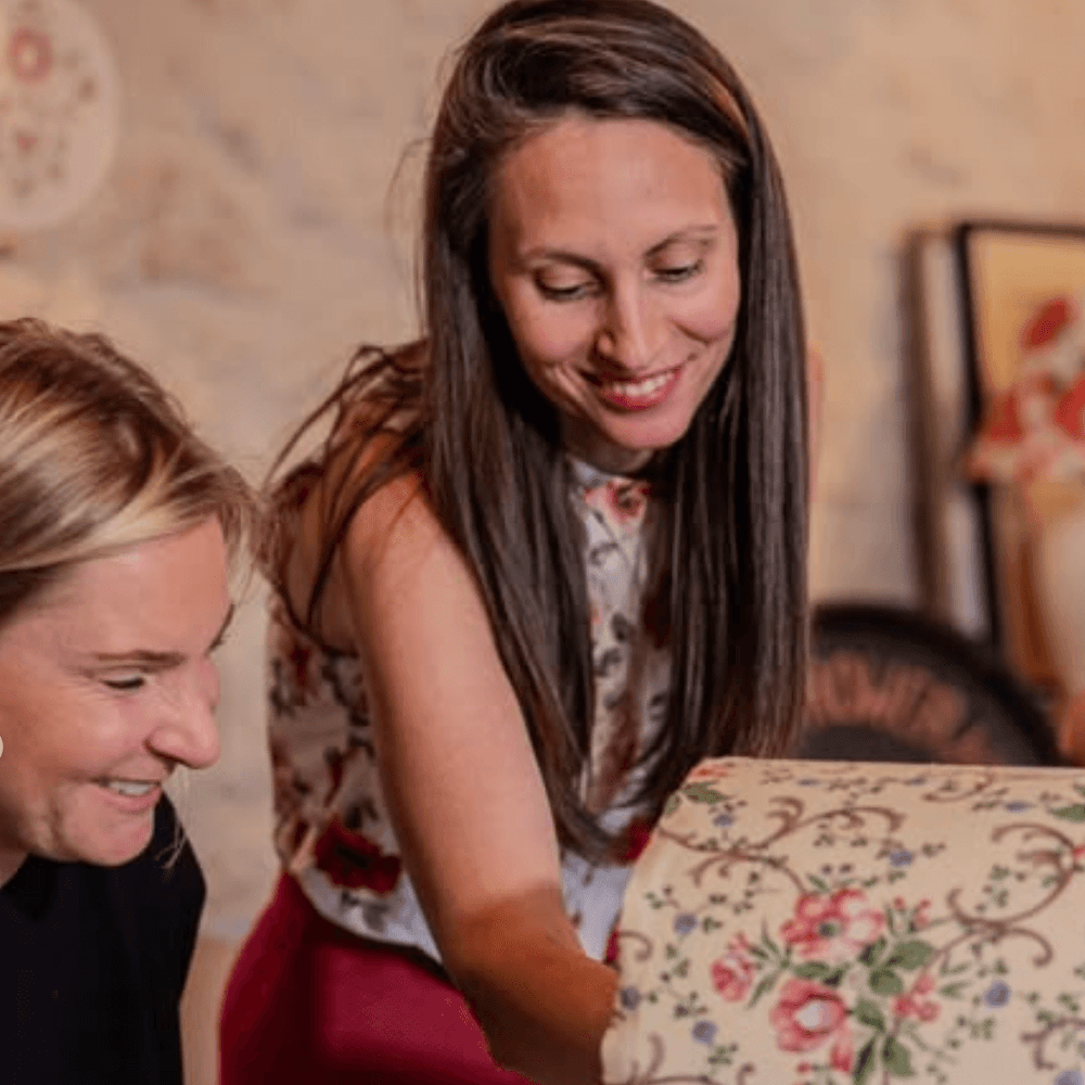 Deux femmes souriantes découvrent ensemble un abat-jour artisanal en tissu fleuri, dans l’univers chaleureux de La Grange des Loupiotes.