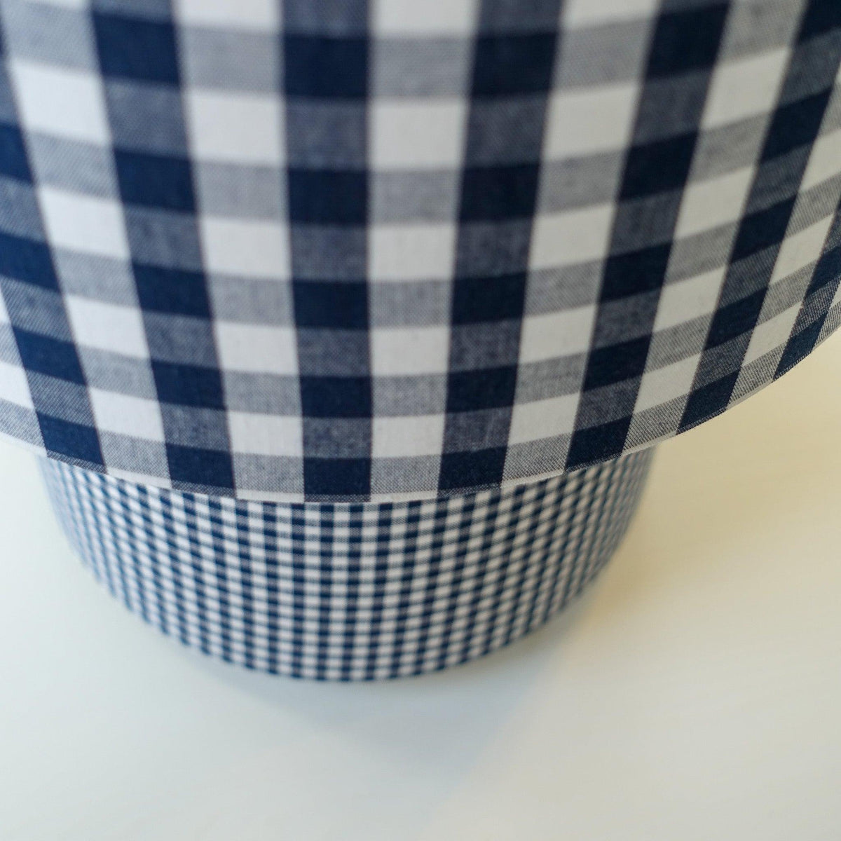 Gros plan sur une lampe habillée de deux motifs vichy bleu et blanc, avec de grands carreaux sur l’abat-jour et un motif plus fin sur le pied.