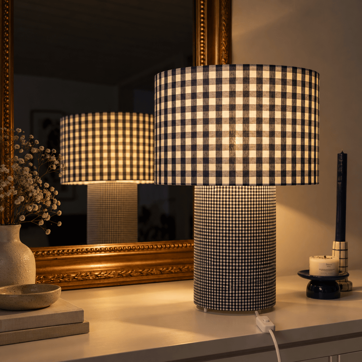Lampe à poser en tissu vichy bleu et blanc, allumée devant un grand miroir doré, avec un abat-jour à grands carreaux et un pied cylindrique à motif plus fin.