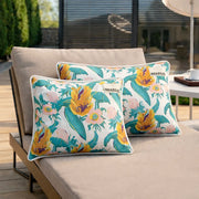 Deux chaises longues beiges avec des coussins floraux colorés, avec des fleurs jaunes et des feuilles vertes, sont installées sur une terrasse en bois à l'extérieur. Une tasse et une soucoupe sont posées sur une table d'appoint à l'arrière-plan.