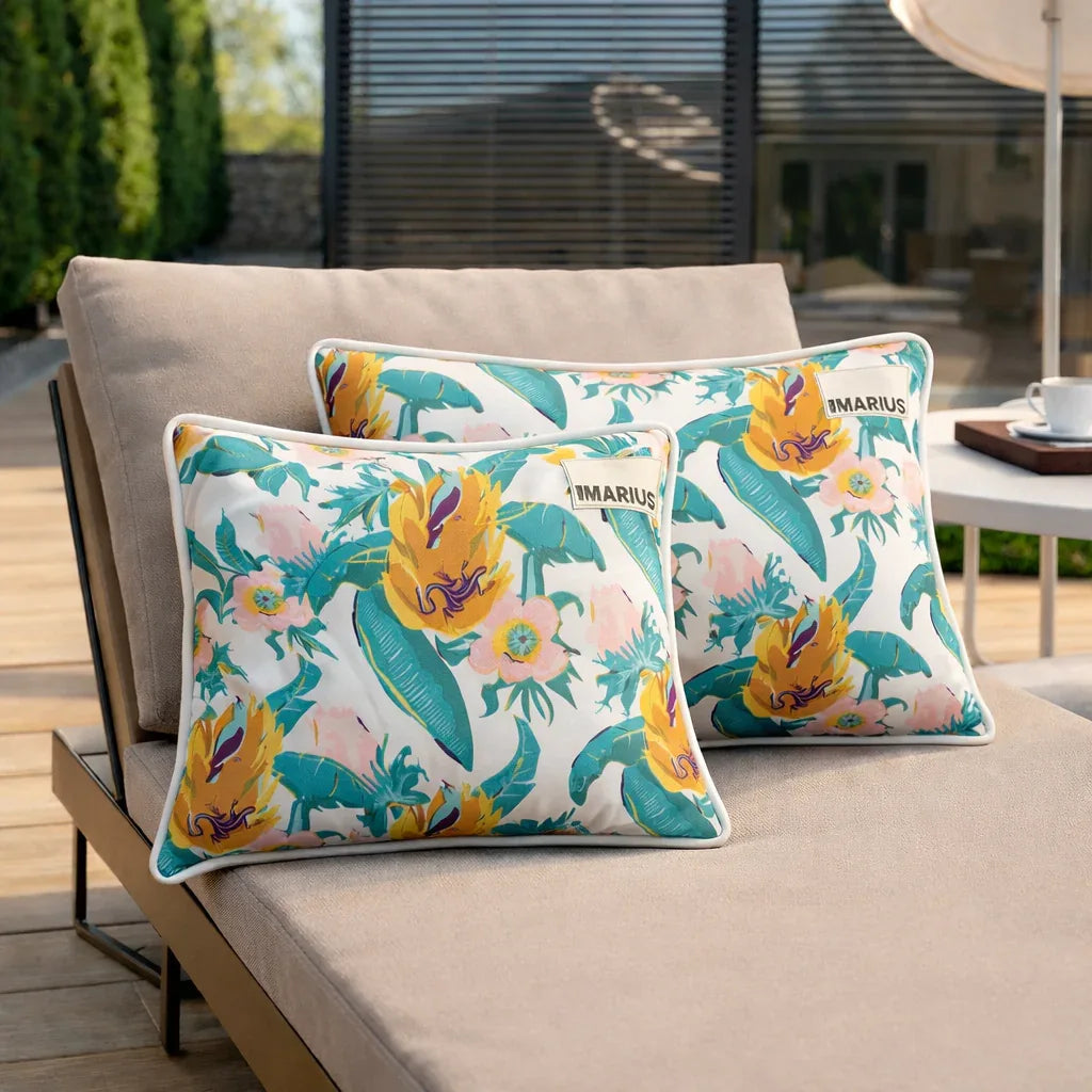 Deux chaises longues beiges avec des coussins floraux colorés, avec des fleurs jaunes et des feuilles vertes, sont installées sur une terrasse en bois à l'extérieur. Une tasse et une soucoupe sont posées sur une table d'appoint à l'arrière-plan.