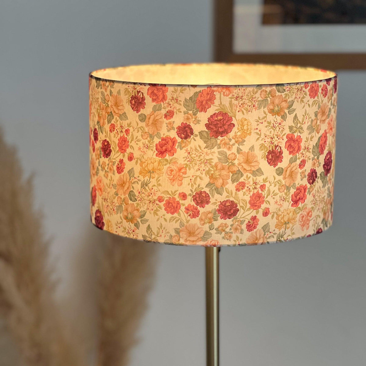 Abat-jour cylindrique fleuri allumé, diffusant une lumière chaude à travers un tissu aux motifs floraux rose, bordeaux, beige et vert, sur un pied en métal doré.