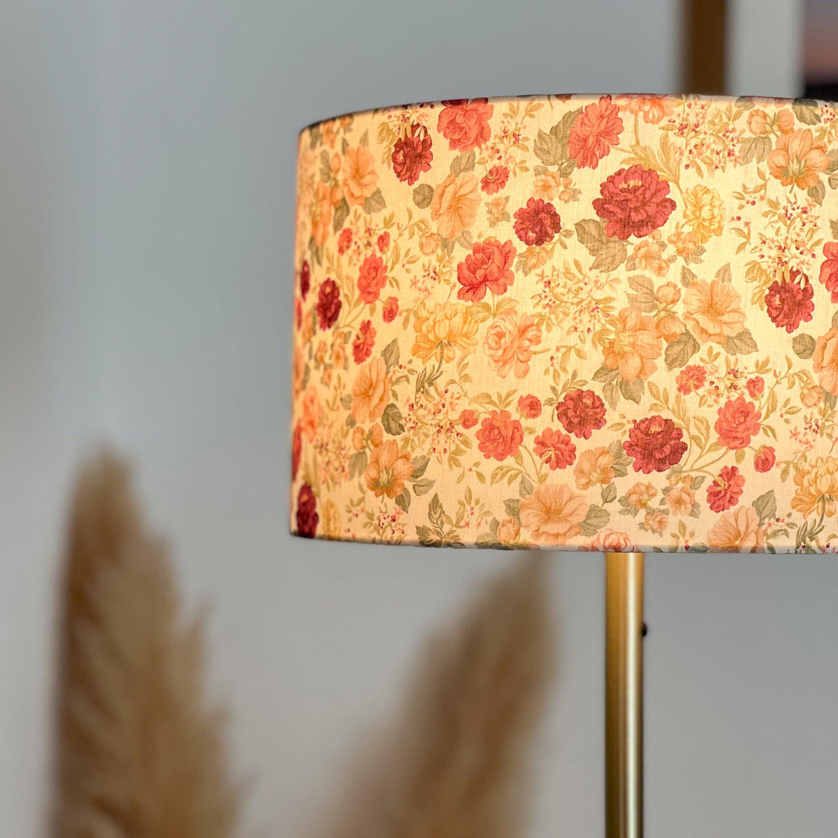 Abat-jour cylindrique fleuri allumé, diffusant une lumière chaude à travers un tissu aux motifs floraux rose, bordeaux, beige et vert, sur un pied en métal doré.