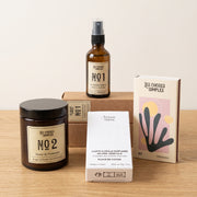 Bougie parfumée en verre ambré, spray d’ambiance 50 ml, carte florale parfumée dans sa boîte et boîte d’allumettes sur une table en bois clair