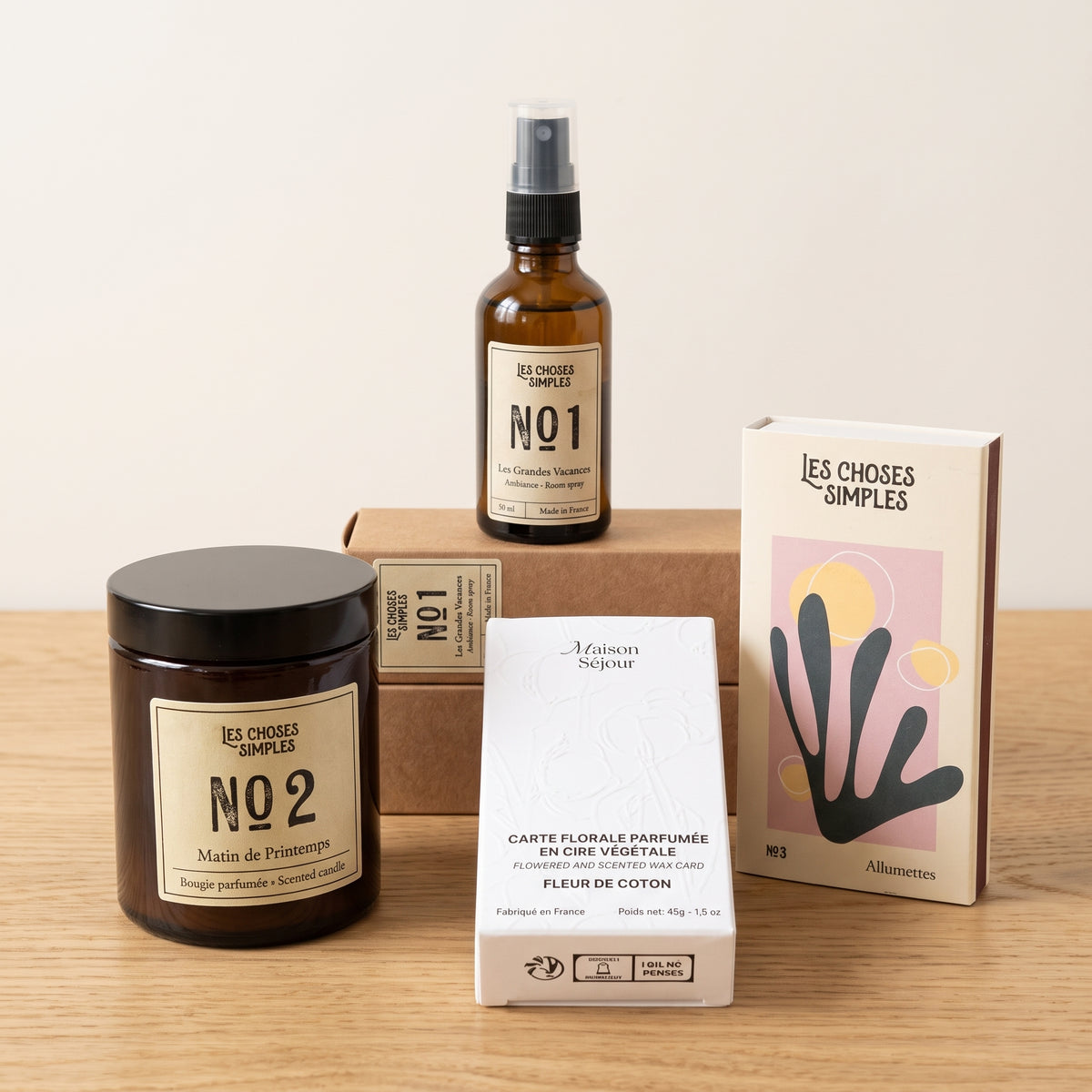 Bougie parfumée en verre ambré, spray d’ambiance 50 ml, carte florale parfumée dans sa boîte et boîte d’allumettes sur une table en bois clair