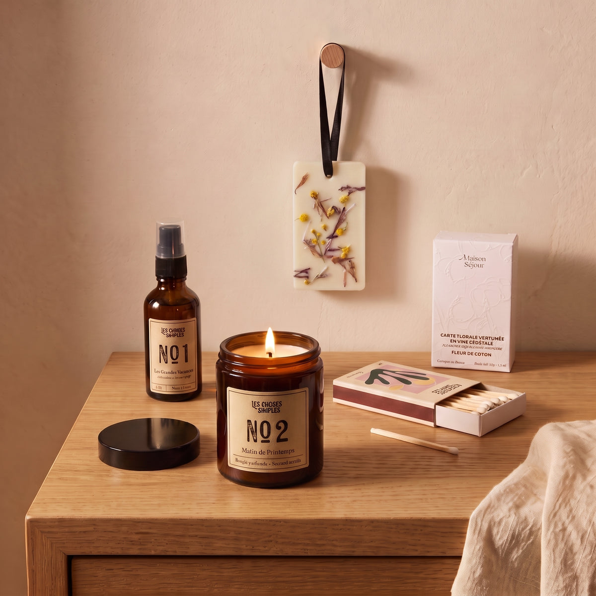 Bougie parfumée allumée sur une table de chevet en chêne, avec le spray d’ambiance, des allumettes et une carte florale suspendue au mur