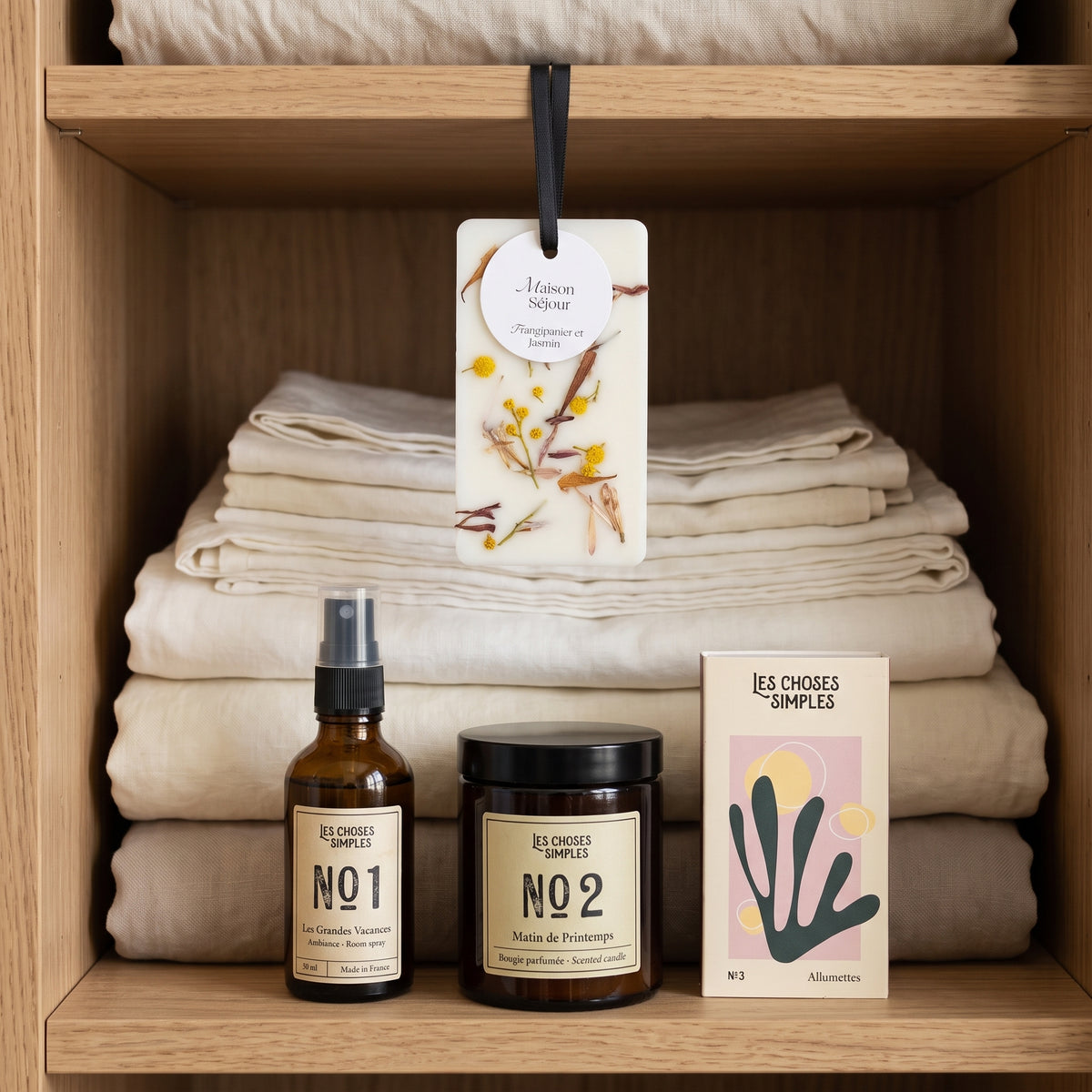 Carte florale parfumée suspendue devant du linge plié dans une armoire en bois, avec le spray d’ambiance, la bougie parfumée et les allumettes