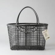 Un panier rectangulaire en plastique noir avec un motif de grille et deux poignées arrondies est posé sur une surface noire assortie. Une étiquette blanche avec un texte et une illustration est attachée à l'une des poignées.