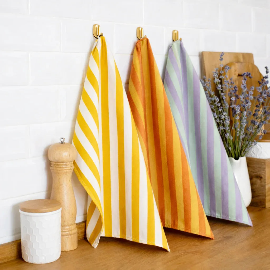 Trois torchons de cuisine rayés jaune, orange et vert sont suspendus à des crochets sur un mur carrelé blanc. À proximité se trouvent un moulin à poivre, une boîte blanche, une planche à découper en bois et un vase avec de la lavande.