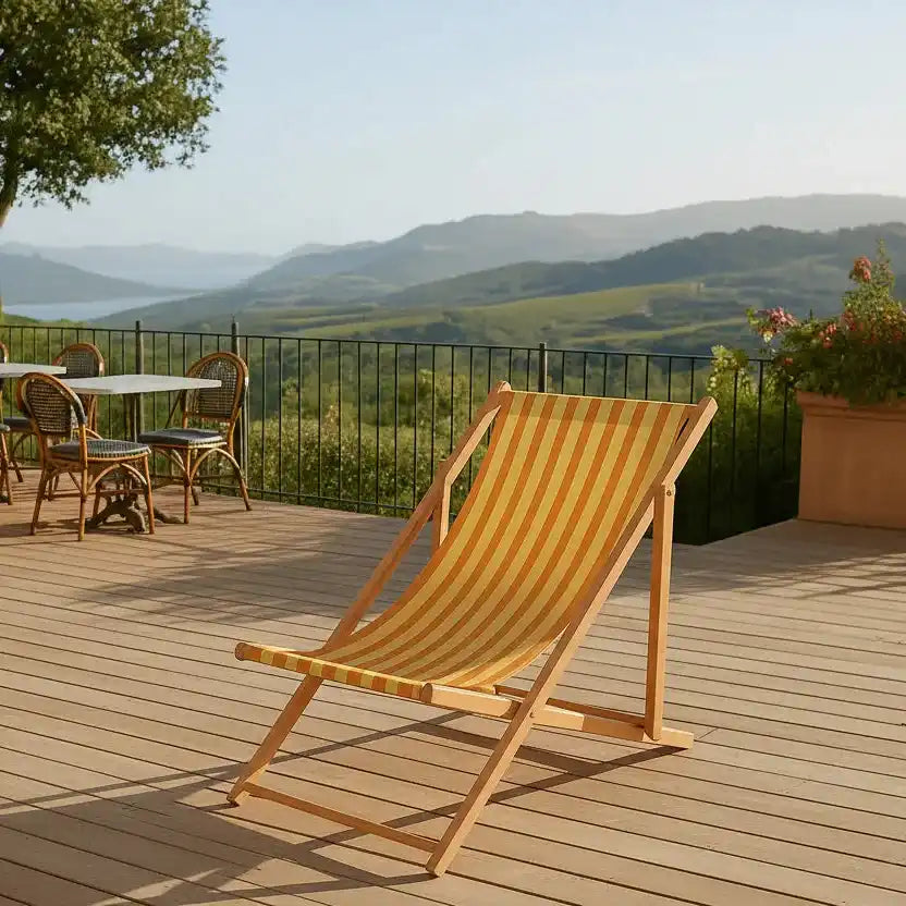 Une chaise longue rayée jaune et orange est assise sur un patio en bois avec une table et des chaises à proximité, surplombant un paysage pittoresque de collines vertes et de montagnes lointaines sous un ciel clair.